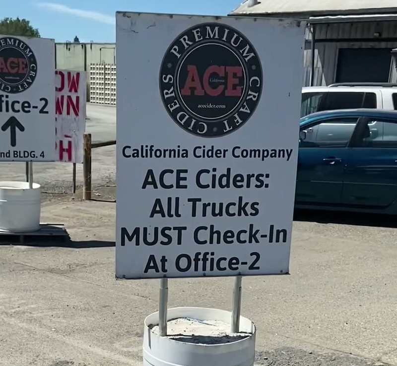 A sign on a gravel yard reading California Cider Company, ACE Ciders: All Trucks MUST Check-In At Office-2, with the ACE roundel above it and warehouse sheds behind.