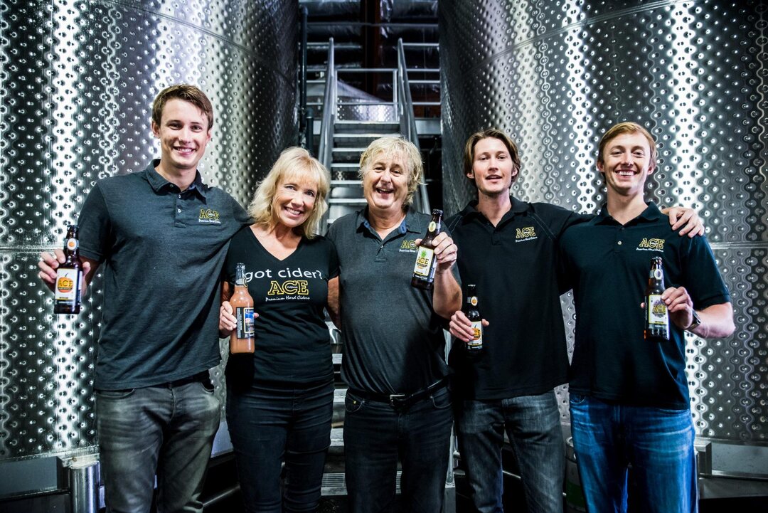 Five people in ACE shirts stand in a row between two tall stainless fermentation tanks, each holding a bottle of cider, with a steel staircase rising behind them.