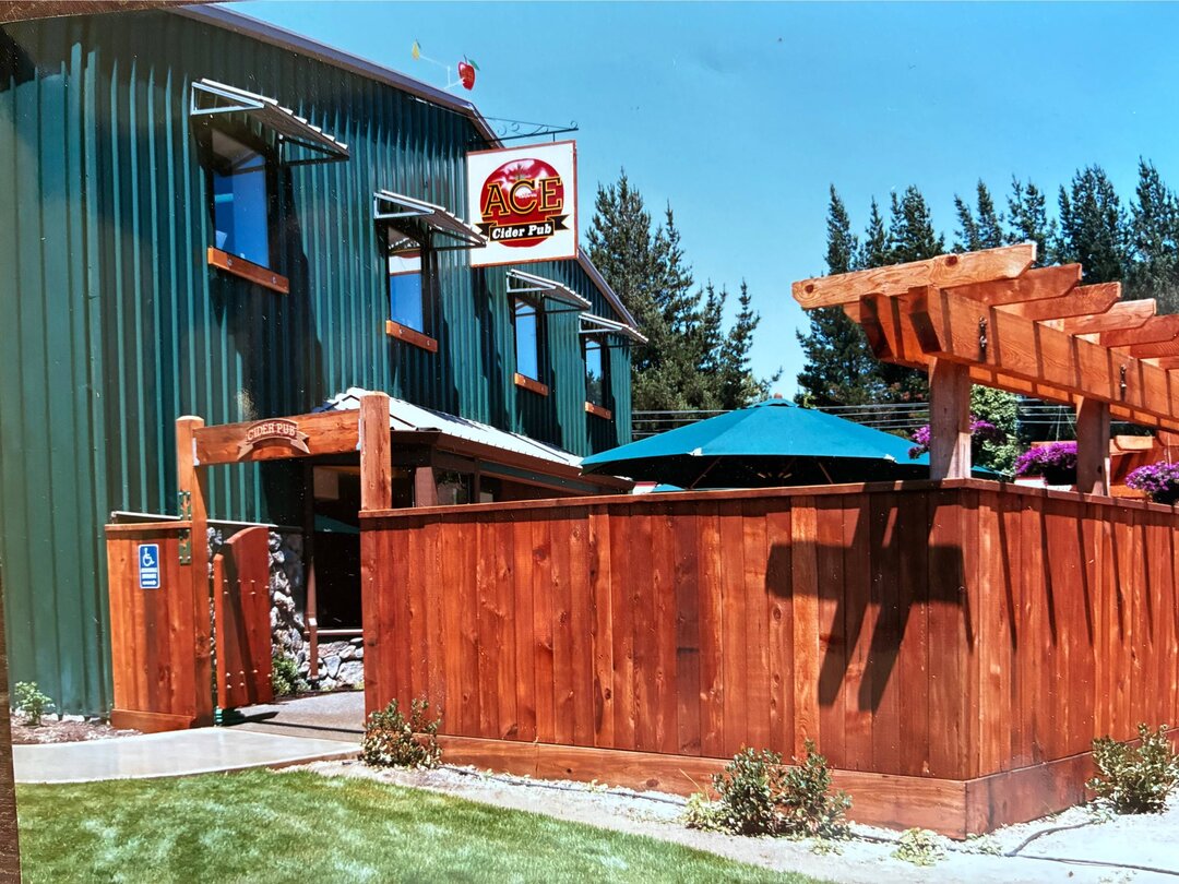 A green corrugated building in sunshine with a hanging sign reading ACE Cider Pub above a carved timber beam reading CIDER PUB, a redwood deck and fence in front, a teal parasol over the garden tables and cypresses behind.