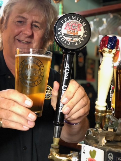 A man behind a bar holding up a glass of cider beside a black ACE Premium Craft Ciders tap handle.