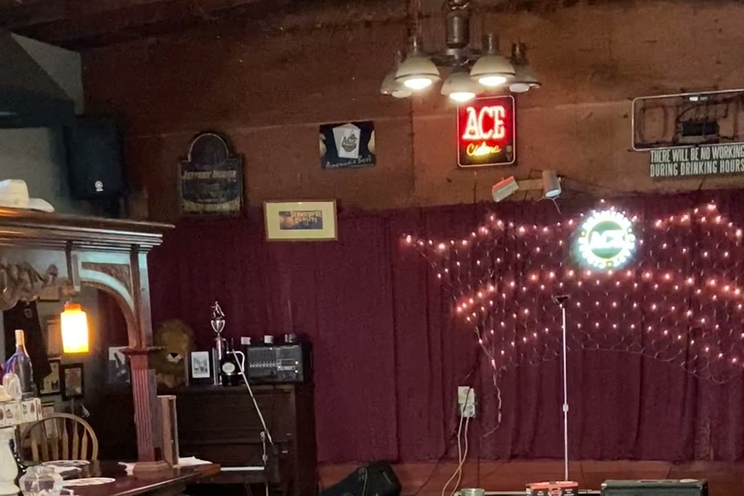 The inside of a timber-ceilinged bar: long communal tables, a small stage with a red curtain and fairy lights, an ACE Ciders neon sign, and cases of cider stacked on a pallet.
