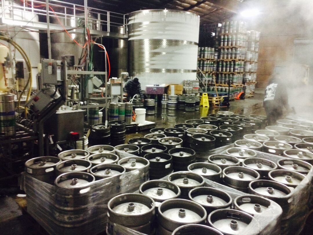 A cidery floor at night: tall stainless fermentation tanks behind, pallets of canned cider stacked to the roof, and dozens of steel kegs filled and wrapped across the foreground, with steam drifting through the light.