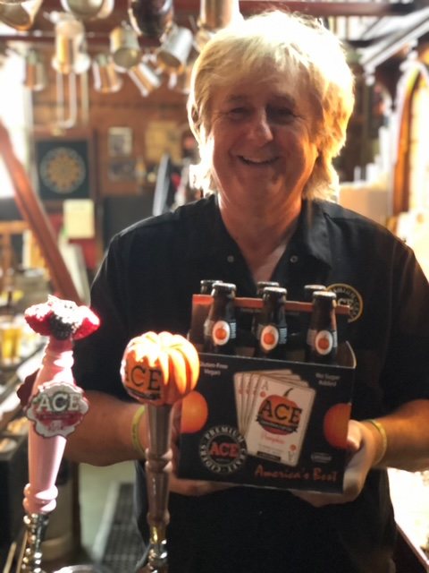 A man behind a bar holding a six-pack of ACE Pumpkin cider, with a pumpkin-topped tap handle and a decorated ACE handle in front of him.