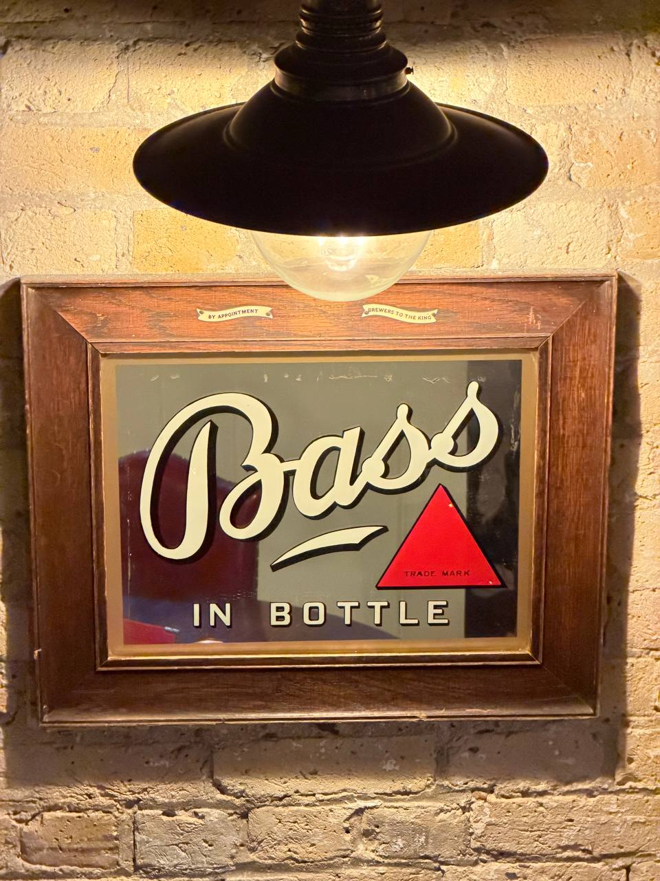 Vintage Bass pub mirror in an oak frame, showing the cursive 'Bass' wordmark next to the red triangle trade mark and the words 'IN BOTTLE'. At the top of the mirror: 'BY APPOINTMENT — BREWERS TO THE KING'. Hung on a whitewashed brick wall under a black industrial pendant light.