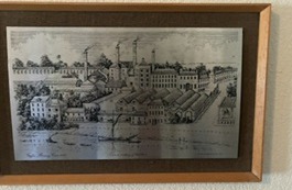 A framed nineteenth-century engraving of a large brewery seen from the river: chimneys and a tall brick block behind rows of pitched roofs, with a barge on the water in the foreground.