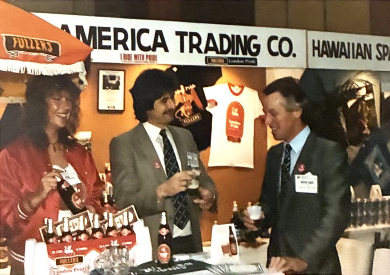 A trade stand under a board reading America Trading Co. A Fuller&rsquo;s pennant hangs at one side and a London Pride T-shirt is pinned up behind. Three people stand at a counter of London Pride six-packs and bottles, two of them holding glasses.